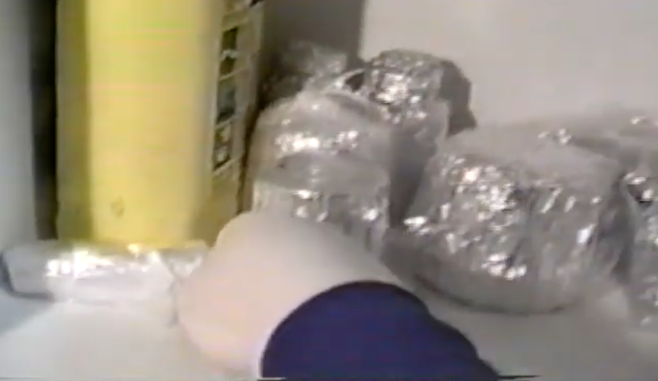 A gloved hand is seen reaching into a fridge containing packages wrapped in foil. A yellow bottle of what appears to be antifreeze is also seen in the shot.